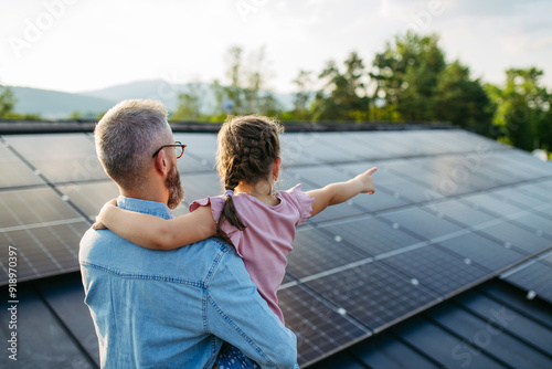 Rear view of dad with girl on roof with solar panels, hugging. Rooftop solar or photovoltaic system. Sustainable future for next generation. photo
