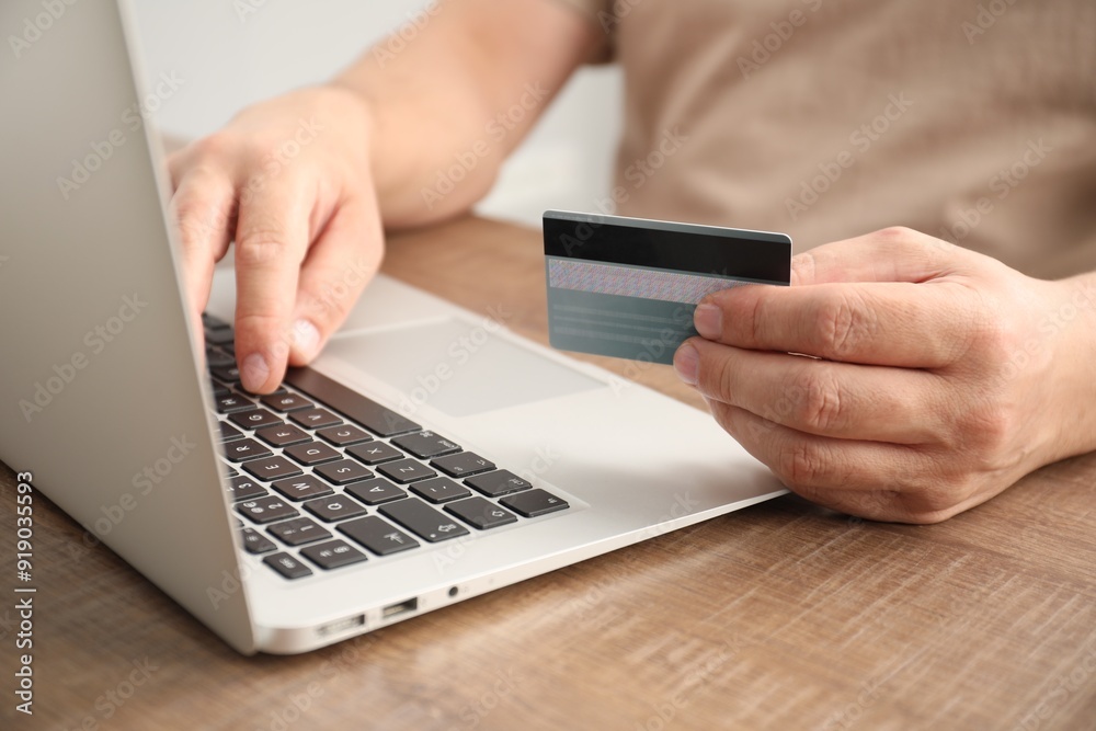 Man with credit card using laptop at wooden table indoors, closeup Man with credit card using laptop at wooden table indoors, closeup
