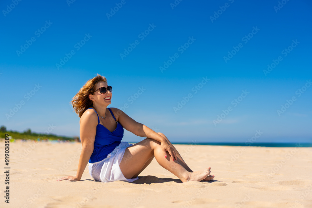 Summer holidays. Beautiful young woman in blue swimsuit and white pareo and sunglasses sitting on ground on sandy beach against backdrop of blue sky. Side view Summer holidays. Beautiful young woman in blue swimsuit and white pareo and sunglasses sitting on ground on sandy beach against backdrop of blue sky. Side view