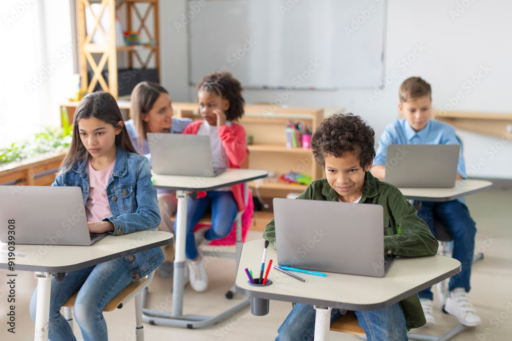 Diverse school kids having lesson, sitting at desks with laptops, studying using digital gadgets. Modern tech elementary education concept Diverse school kids having lesson, sitting at desks with laptops, studying using digital gadgets. Modern tech elementary education concept