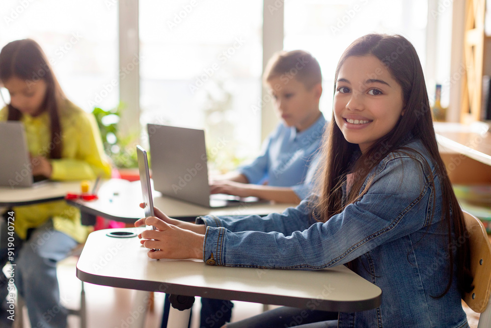 Happy European school girl smiling at camera while studying with laptop computer, sitting at table in classroom , pupils having tech lesson Happy European school girl smiling at camera while studying with laptop computer, sitting at table in classroom , pupils having tech lesson