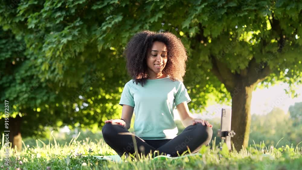 Young happy african american sportswoman practices yoga on a mat in city park using an application on her smartphone. Black woman is sitting in lotus position, and listening to online course on phone Young happy african american sportswoman practices yoga on a mat in city park using an application on her smartphone. Black woman is sitting in lotus position, and listening to online course on phone