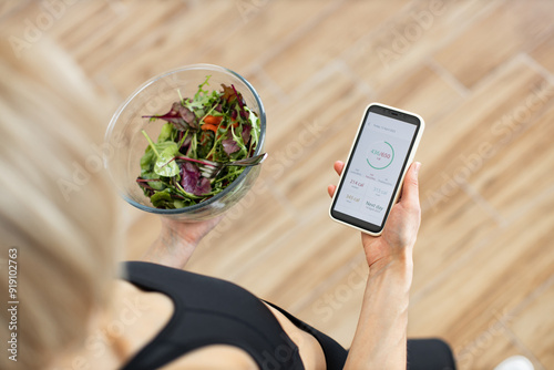 Fotografija Woman in fitness clothing tracking daily calorie intake on smartphone while holding bowl of fresh salad
