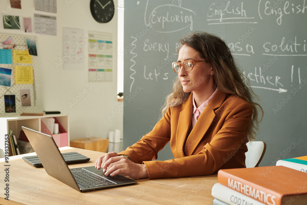 Fototapeta premium Side view portrait of adult woman with long hair using laptop in school classroom with blackboard in background