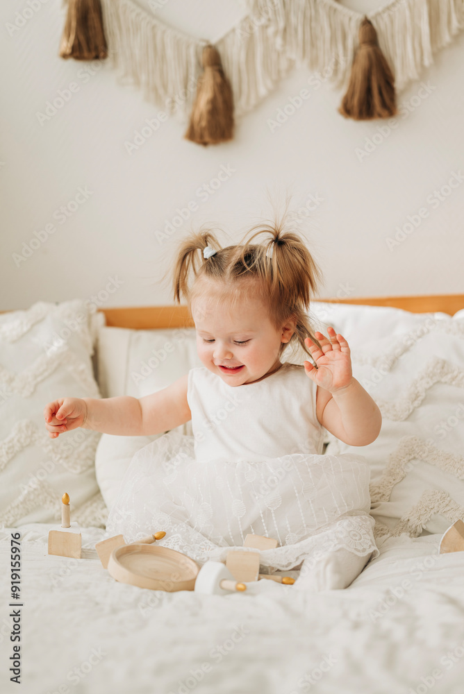 Fototapeta premium Cute beautiful girl baby sitting on the bed in the bedroom and playing in eco wooden toys cubes and pyramid