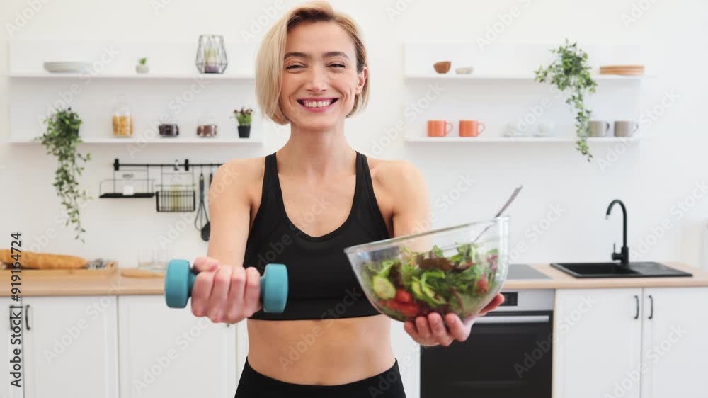 Smiling woman in sportswear holding salad bowl and dumbbell in modern kitchen, representing healthy lifestyle, diet, and fitness. Smiling woman in sportswear holding salad bowl and dumbbell in modern kitchen, representing healthy lifestyle, diet, and fitness.