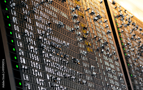 Split flap display at the airport with folding black letter panels showing arrival and departure times and destinations in the moment of scrolling. Analog info medium from below with selective focus.