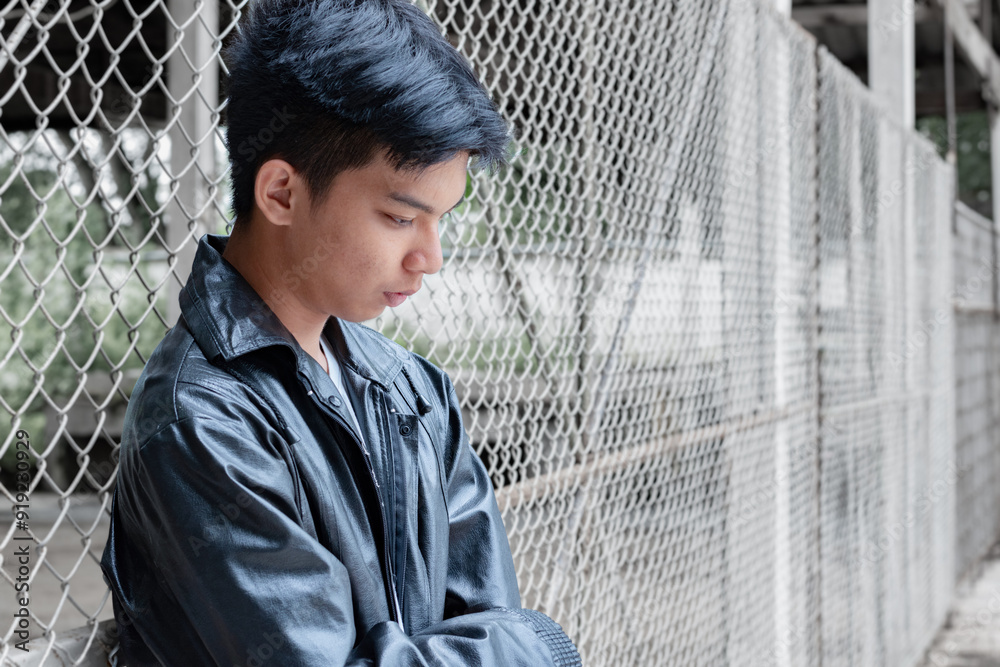 Φωτογραφία Asian teenboy in a black shirt sits pressed against a metal fence panel in a juvenile detention facility lonely, awaiting further release, depression,freedom and detention of young people concept