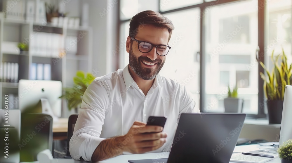 Smiling businessman using phone and laptop in modern office. He is wearing glasses and a white shirt. Commercial use Smiling businessman using phone and laptop in modern office. He is wearing glasses and a white shirt. Commercial use