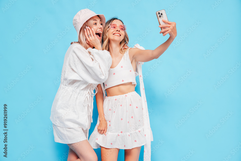 Two young beautiful smiling blond hipster female in trendy summer clothes. Blonde carefree women posing near blue wall. Positive models having fun. Cheerful and happy, panama hat, take selfie Two young beautiful smiling blond hipster female in trendy summer clothes. Blonde carefree women posing near blue wall. Positive models having fun. Cheerful and happy, panama hat, take selfie