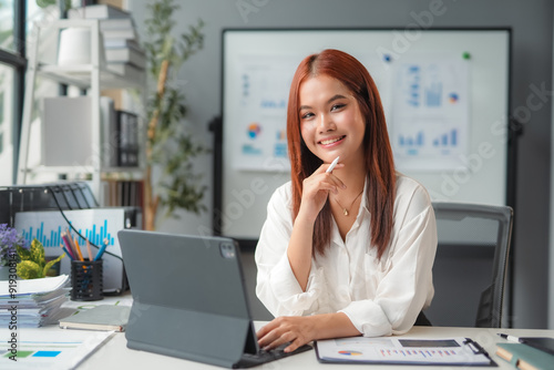 Young asia businesswoman holding pen and using tablet, looking at camera with smile while sitting at office desk