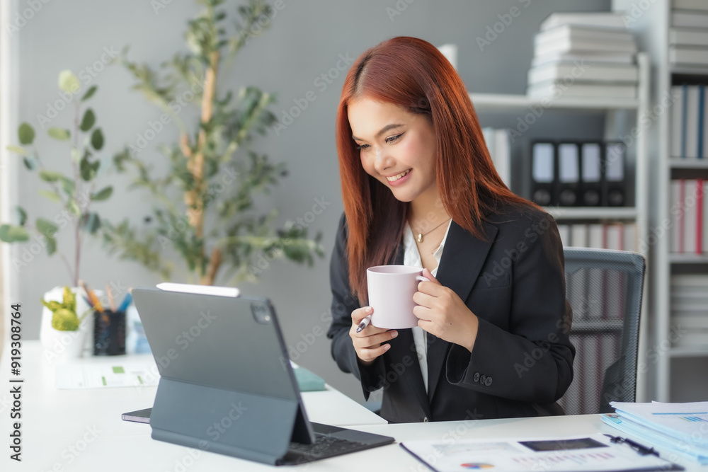 Asian businesswoman smiles while enjoying coffee at work in a modern office, focused on her laptop amidst technology and paperwork, exuding confidence and professionalism Asian businesswoman smiles while enjoying coffee at work in a modern office, focused on her laptop amidst technology and paperwork, exuding confidence and professionalism
