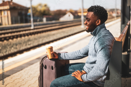 Happy man with suitcase enjoys drinking coffee while sitting on a bench at the railway station.	