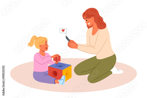 Happy mother takes photo of little daughter playing with blocks on mobile phone camera. Mother rejoices at success of pre-teen child demonstrating talent and readiness for development