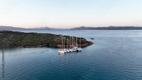 Seven sailboats moored close to the rocky shore of a small island in the Mediterranean Sea, footage being taken at sunset in Sardinia.