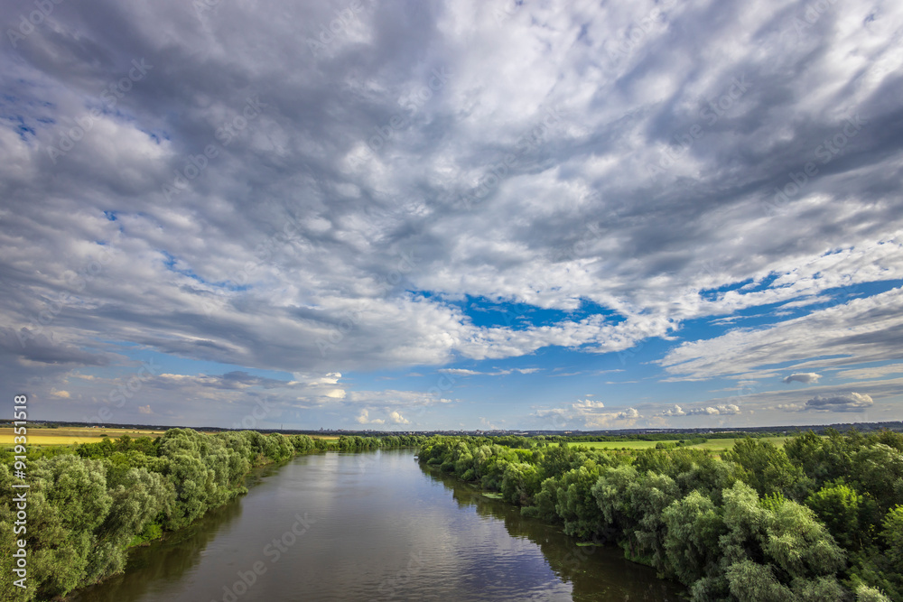A peaceful river flows through vibrant green banks, reflecting an expansive sky filled with dynamic clouds as the day transitions to evening. A peaceful river flows through vibrant green banks, reflecting an expansive sky filled with dynamic clouds as the day transitions to evening.