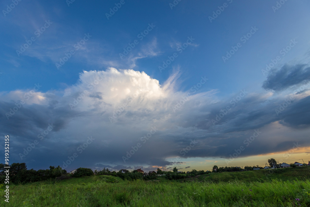 An expansive, white cumulus cloud dominates the sky above a field of green grass, with dark storm clouds gathering in the distance. The evening light creates a soft glow on the horizon. An expansive, white cumulus cloud dominates the sky above a field of green grass, with dark storm clouds gathering in the distance. The evening light creates a soft glow on the horizon.