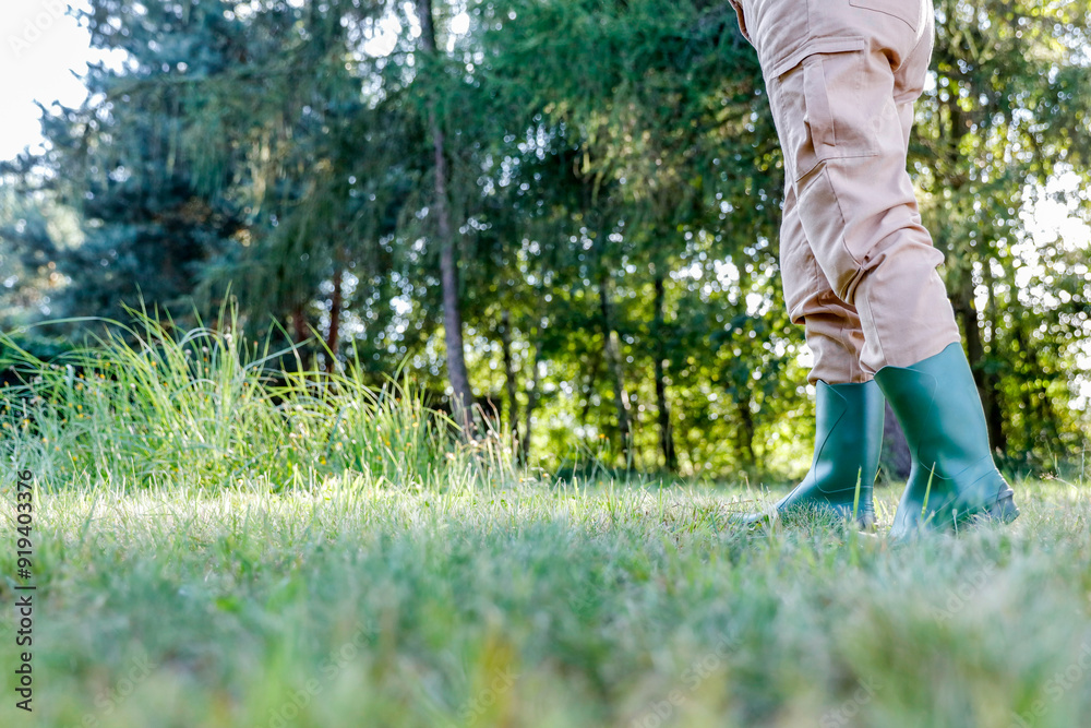 Woman gardener in brown garden overalls and rubber boots on the garden in warm autumn sunny day. Lush vegetation and grass and trees background. Autumn gardening. Woman gardener in brown garden overalls and rubber boots on the garden in warm autumn sunny day. Lush vegetation and grass and trees background. Autumn gardening.