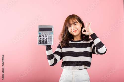 Tax day concept. Woman confident smiling holding electronic calculator and show OK gesture, excited happy Asian female studio shot isolated on pink background, Account and finance counting income