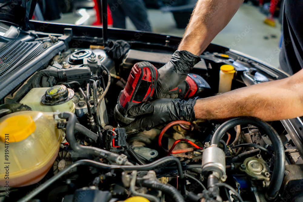 Obraz premium close up in a car service a master in a black T-shirt uses a drill to unscrew the bolts in the engine part of the car replacing spare parts