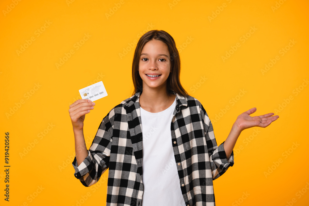 Little kid teenager child school girl wears casual clothes hold in hand mock up of credit bank card isolated on bright orange yellow background. Childhood banking concept Little kid teenager child school girl wears casual clothes hold in hand mock up of credit bank card isolated on bright orange yellow background. Childhood banking concept