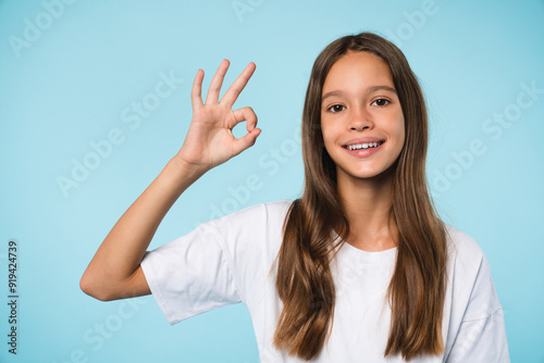 Smiling with toothy smile caucasian teenage girl schoolchild pupil student showing okay gesture close up portrait isolated in blue background