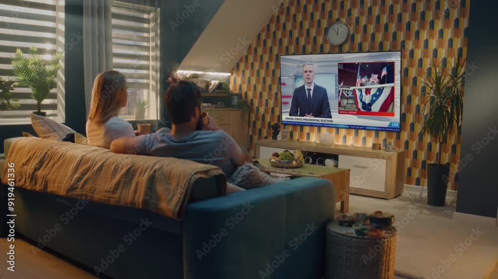 Caucasian couple watches news broadcast about US presidential elections on the big TV display, talks and drinks tea sitting on couch at home. Breaking news with male newscaster displayed on TV screen.