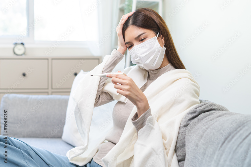 Sick, influenza asian young woman wearing face mask, have fever hand touching forehead, holding thermometer for check measure body temperature, illness while sitting rest on sofa at home. Health care. Sick, influenza asian young woman wearing face mask, have fever hand touching forehead, holding thermometer for check measure body temperature, illness while sitting rest on sofa at home. Health care.