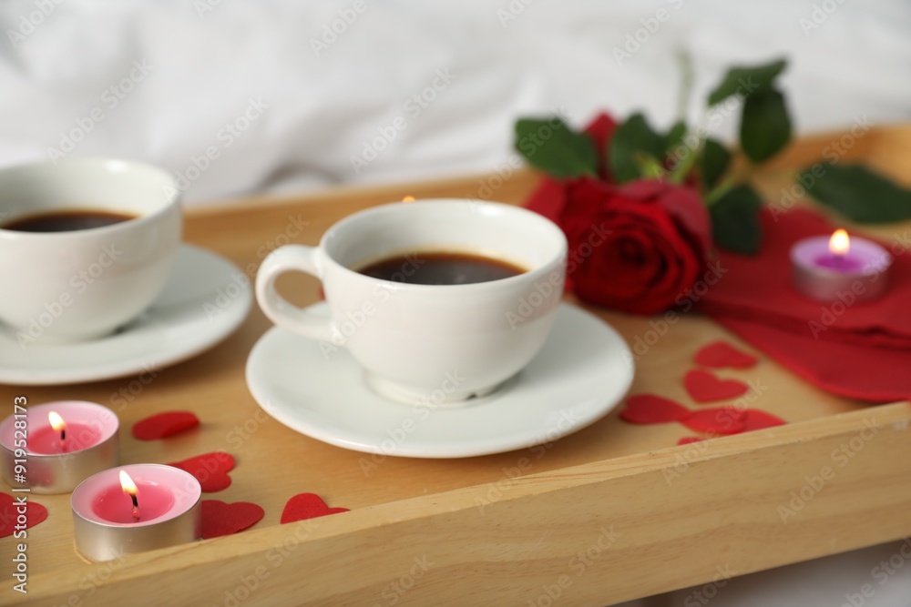 Wooden tray with burning candles, rose, cups of coffee and red paper hearts on bed indoors, closeup