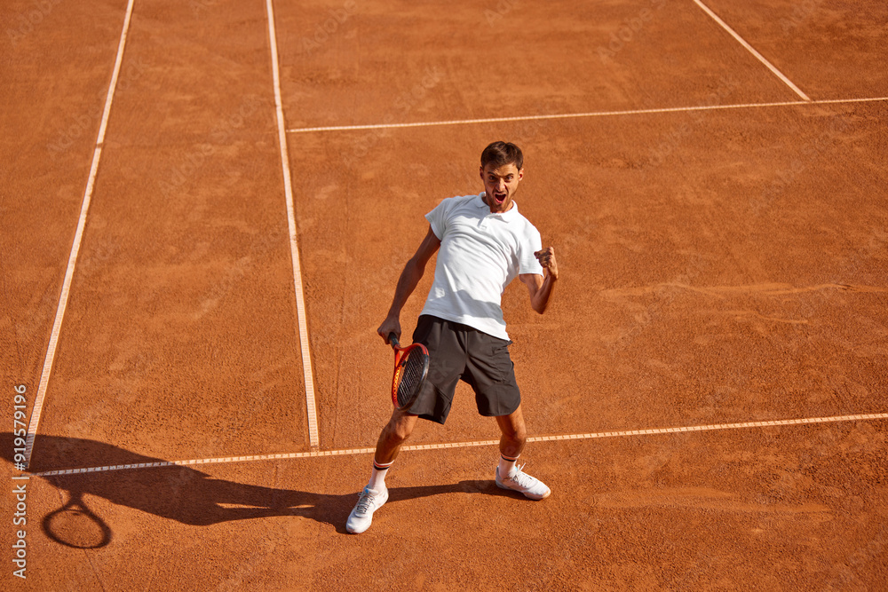 Emotional young man, tennis player on outdoor clay court, showing happiness of successful game. Champion, winner. Victory emotions. Concept of sport, competition, active and healthy lifestyle Emotional young man, tennis player on outdoor clay court, showing happiness of successful game. Champion, winner. Victory emotions. Concept of sport, competition, active and healthy lifestyle