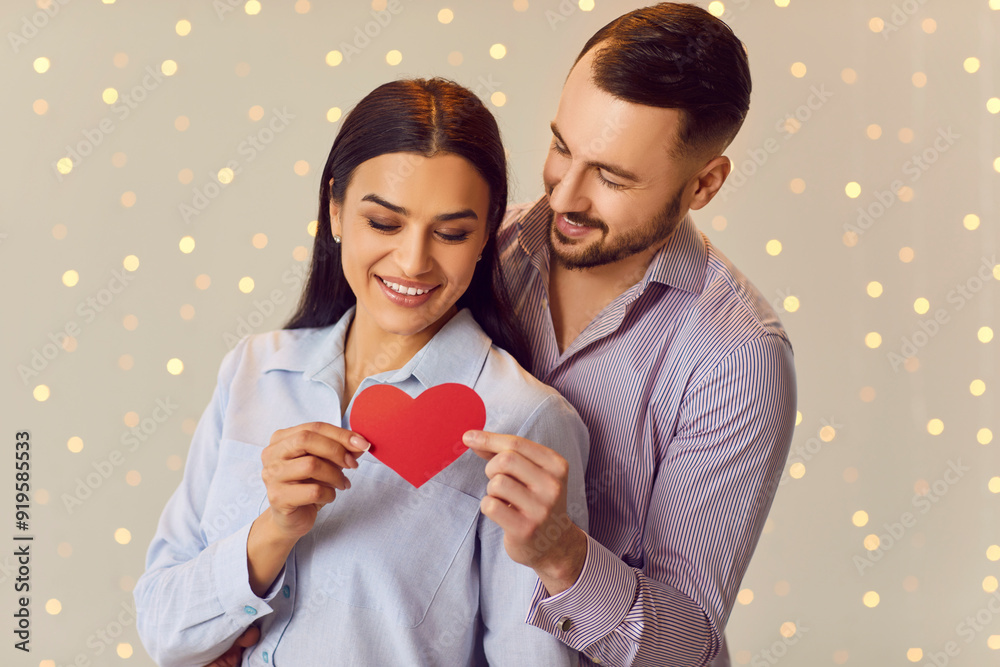 Portrait of a young happy smiling man holding red paper heart in hand and hugging brunette girl standing indoors on lights bokeh background. Happy couple in love. Valentines day concept. Portrait of a young happy smiling man holding red paper heart in hand and hugging brunette girl standing indoors on lights bokeh background. Happy couple in love. Valentines day concept.