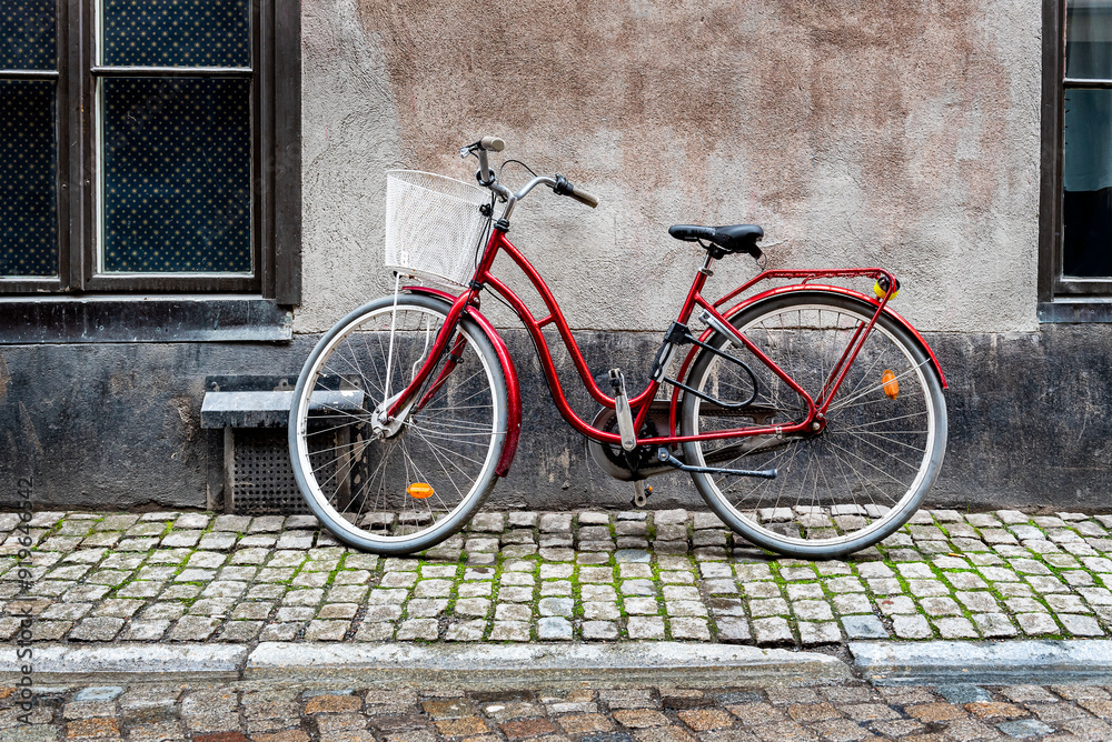 Red lady's bicycle with a shopping basket in front of a blue-grey building wall on a cobblestone street in a vintage look. Red lady's bicycle with a shopping basket in front of a blue-grey building wall on a cobblestone street in a vintage look.