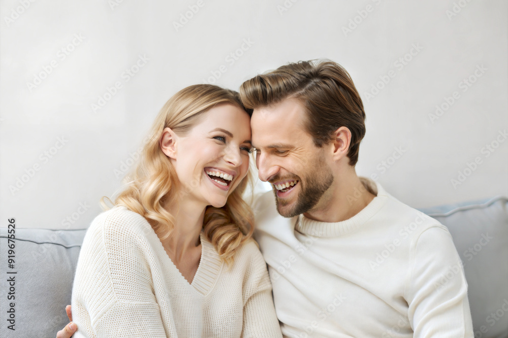 Laughing couple sitting close on a sofa, happy and relaxed, wearing light sweaters against a light wall Laughing couple sitting close on a sofa, happy and relaxed, wearing light sweaters against a light wall