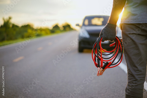 Close-up of a man's hand holding a car battery jumper cable with a dead car battery on the roadside. Repair and maintenance concepts