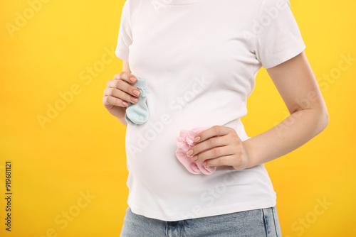 Expecting twins. Pregnant woman holding two pairs of socks on yellow background, closeup