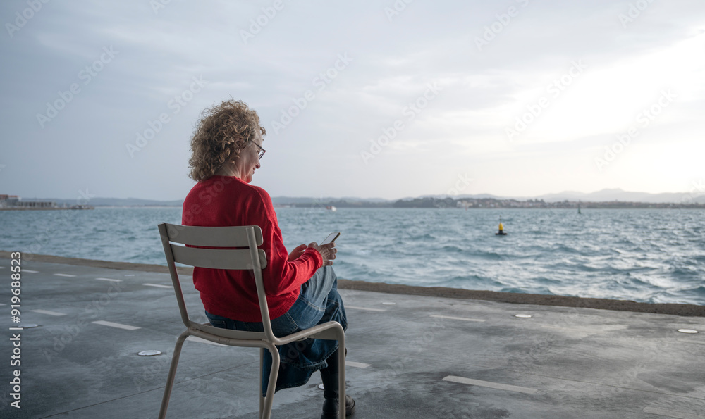 Fototapeta premium Woman sitting on a chair by the sea, using a smartphone, with a distant shoreline in view
