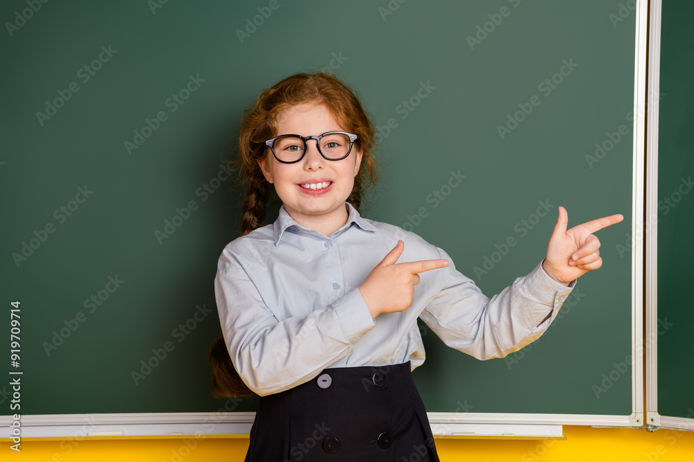Photo of small schoolkid girl indicate fingers empty space wear uniform isolated on yellow color blackboard background
