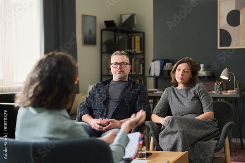 Couple sitting in office discussing relationship issues with therapist holding clipboard. Room has bookshelf, lamp, abstract painting, and large window providing natural light photo