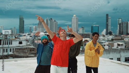 Multicultural teenagers dancing while looking at camera on rooftop with urban city view. Professional break dance group moving to hip-hop music with friends cheer. Outdoor sport 2024. Hiphop.