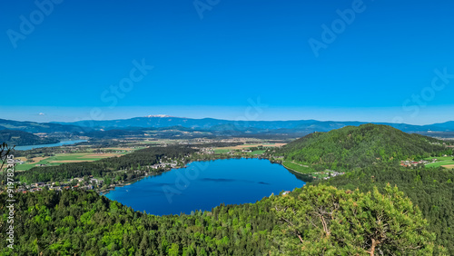 Breathtaking aerial view of a tranquil lake Klopein nestled amidst lush green forest in Carinthia, Austria. Crystal-clear water reflects vibrant blue sky. Rolling hills and mountains in Austrian Alps