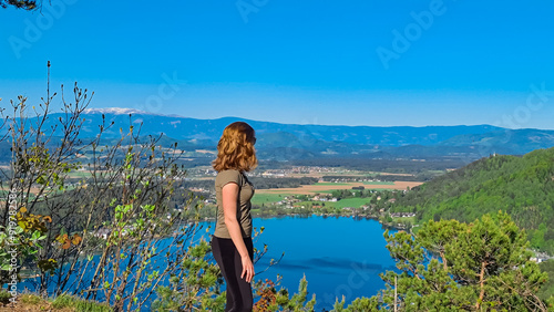 Woman at Kitzelberg with aerial view of lake Klopein nestled amidst lush green forest in Carinthia, Austria. Crystal-clear water reflects vibrant blue sky. Rolling hills and mountains in Austrian Alps photo