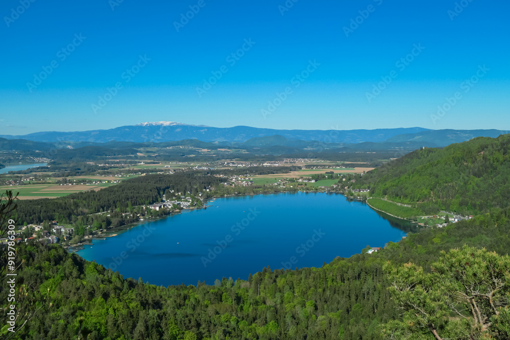 Fototapeta premium Breathtaking aerial view of a tranquil lake Klopein nestled amidst lush green forest in Carinthia, Austria. Crystal-clear water reflects vibrant blue sky. Rolling hills and mountains in Austrian Alps