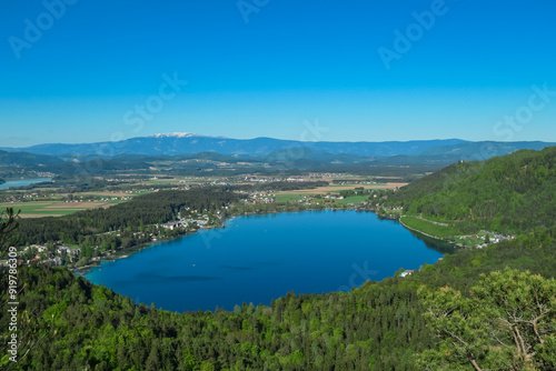 Breathtaking aerial view of a tranquil lake Klopein nestled amidst lush green forest in Carinthia, Austria. Crystal-clear water reflects vibrant blue sky. Rolling hills and mountains in Austrian Alps