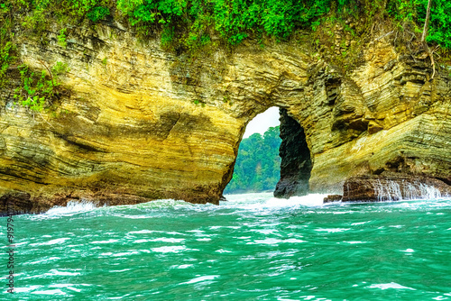 Natural rock arch formation along the Pacific coast, with lush green vegetation atop and turquoise water below, leading to a view of the forested landscape beyond. High-quality photo. Uvita Puntarenas photo