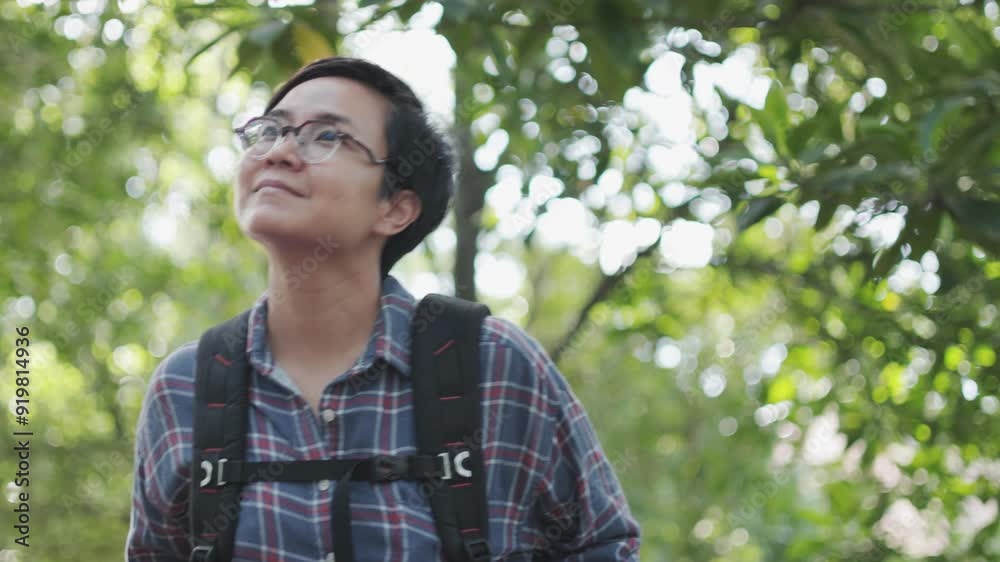 A young, confident Asian woman with short hair is walking through a lush forest, wearing a plaid shirt and carrying a backpack, and enjoying the serenity and greenery of the nature outdoors. A young, confident Asian woman with short hair is walking through a lush forest, wearing a plaid shirt and carrying a backpack, and enjoying the serenity and greenery of the nature outdoors.