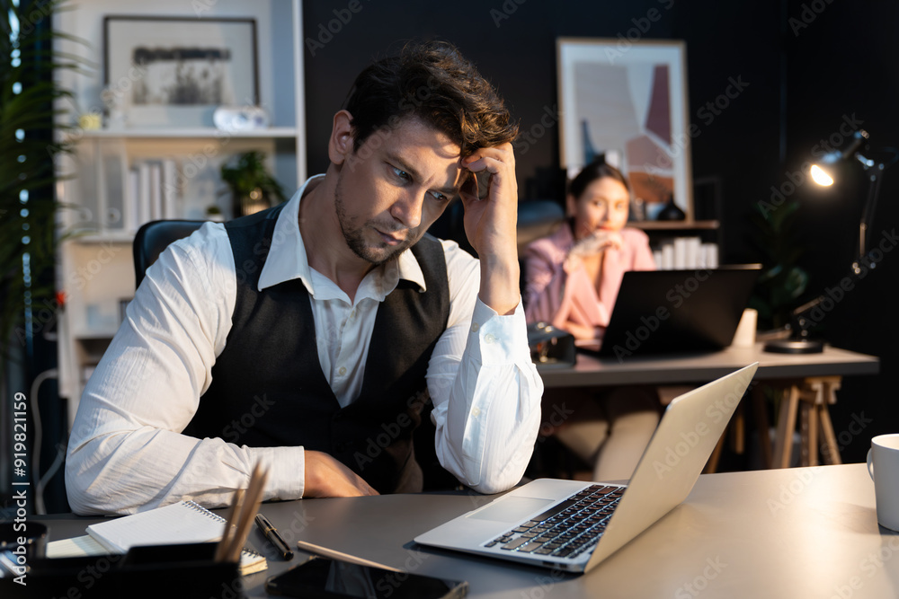 Sitting man with serious face thinking creative business project planning at night lighting time at modern office with blurry secretary checking paper report with folder background concept. Postulate. Sitting man with serious face thinking creative business project planning at night lighting time at modern office with blurry secretary checking paper report with folder background concept. Postulate.