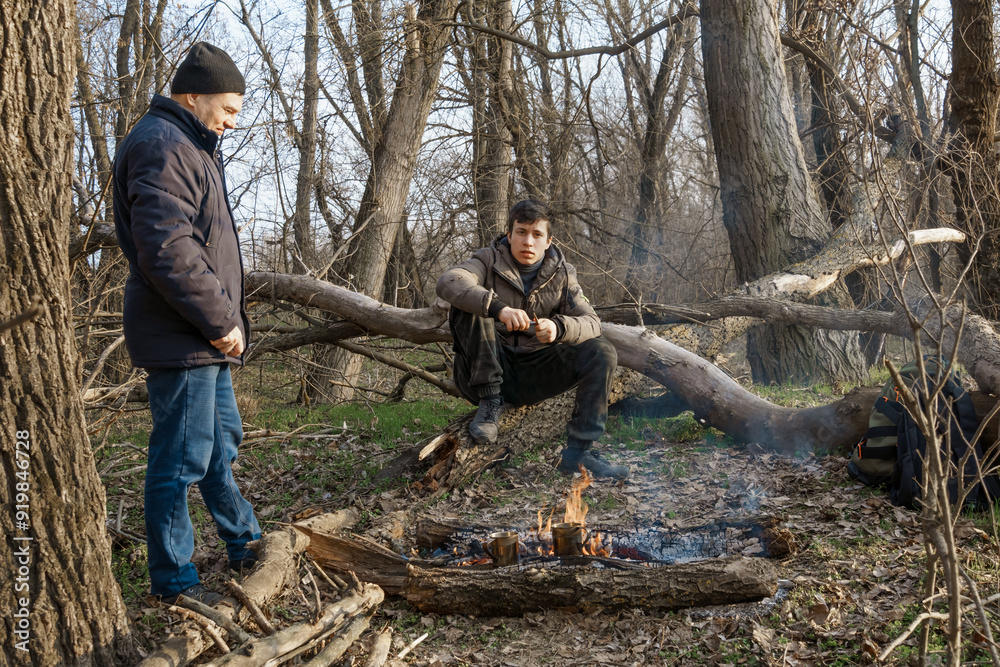 Two men make a campfire for cooking in the forest, father and son sitting on a log while hiking and outdoor activities, early spring landscape Two men make a campfire for cooking in the forest, father and son sitting on a log while hiking and outdoor activities, early spring landscape