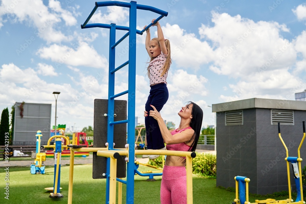 Fototapeta premium Mother assisting daughter on playground equipment at a sunny park in the afternoon
