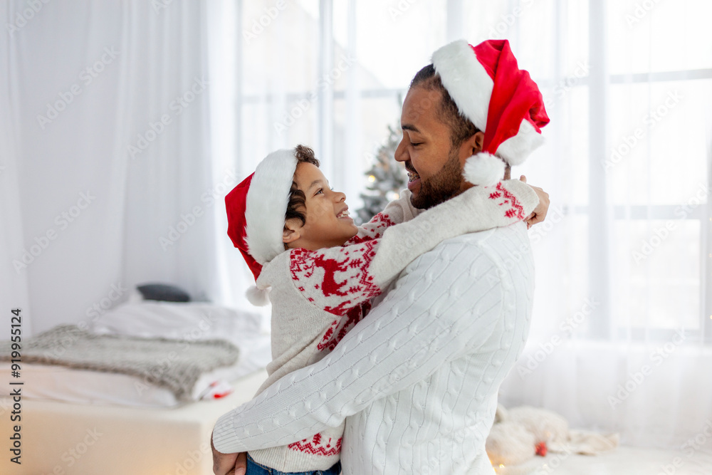 Fototapeta premium african american teenage boy in santa hat hugs dad at home for christmas, man wishes son happy new year and celebrates together