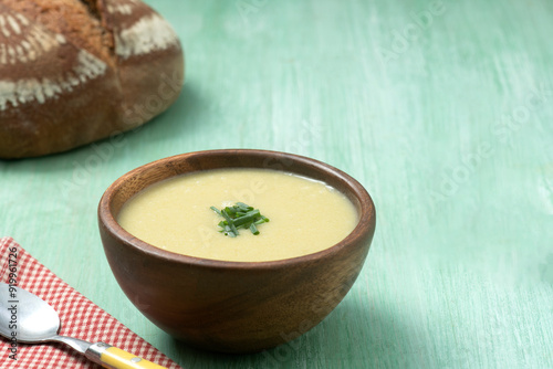 A yellow vegetable soup decorated with chives in a large carved wooden bowl on a green vintage table with red checkered napkin and spoon and rustic sour dough loaf of bread in the back